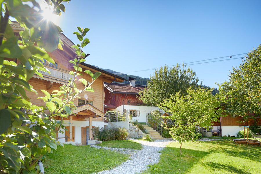 Holzchalets mit blühendem Garten, Bäumen und einer Berglandschaft im Hintergrund, erhellt von sommerlicher Sonne.